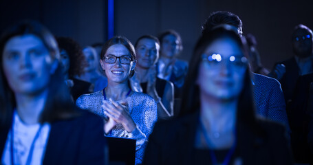 Beautiful Female Sitting in a Dark Crowded Auditorium at a Conference. Young Woman Clapping After a Successful Keynote Presentation. Specialist Inspired by Latest Technology. - obrazy, fototapety, plakaty