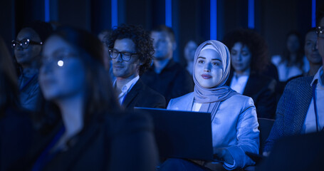 Arab Female Sitting in a Dark Crowded Auditorium at a Tech Conference. Young Muslim Woman Using...