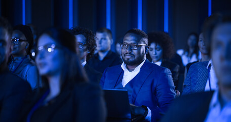 African Male Sitting in a Dark Crowded Auditorium at a Tech Conference. Professional Using a Laptop Computer. Specialist Watching Innovative Technology Presentation About New Software Solutions.