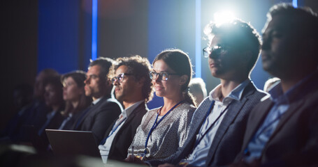 Young Woman Sitting in Crowded Audience at a Business Conference. Female Delegate Smiling and Using Laptop Computer. Manager Watching Motivational Presentation About Angel Investing.