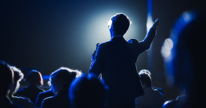 Backview Of A Stylish Young Businessman In A Dark Crowded Auditorium At A Startup Summit. Young Man Talking To A Microphone During A Q And A Session. Entrepreneur Happy With Event Speaker.