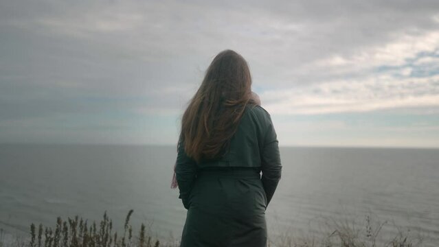 Back view of long haired brunette woman standing in green coat on a cliff and looking to a sea view on moody cloudy day. Thoughtful lonely female overthinking near ocean. Wind blows airy hair.
