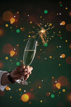 A male hand in white shirt holding glass of sparkling wine champagne with sparkler in it on dark green background with bokeh and christmas lights