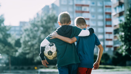 Two Young Caucasian Boys Embracing Each Other while Walking in Neighborhood Football Pitch. Soccer Players Carrying a Ball. Concept of Sports, Childhood, Friendship. Cold Desaturated Color Grading.