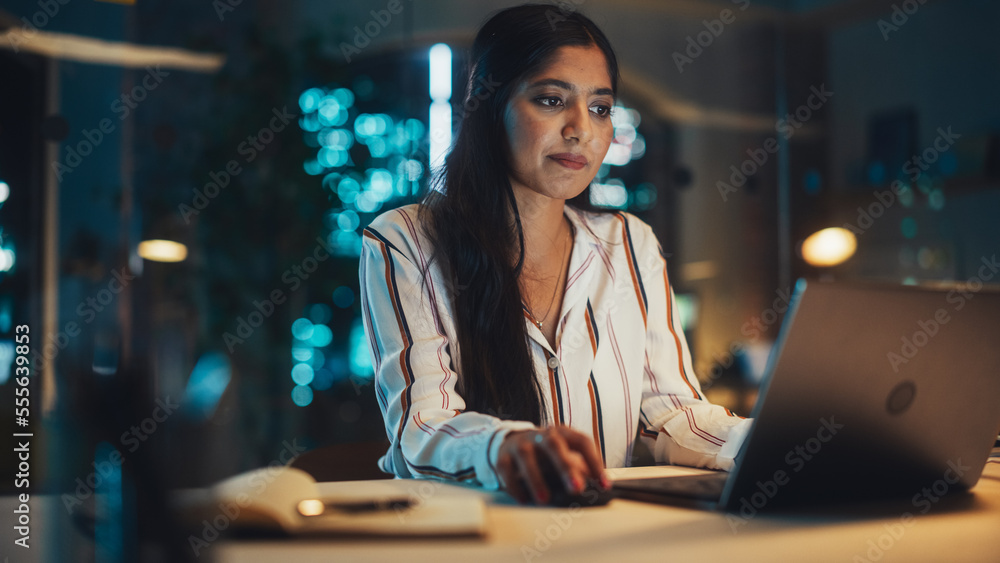 Poster portrait of a beautiful indian female working on laptop computer in a company office at night. young