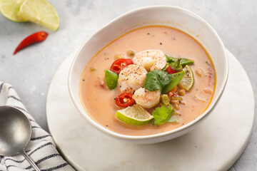 a spicy thai coconut soup or curry made of coconut milk, cherry tomatoes, snow pea, chili pepper, lime, ginger, shrimps and fresh cilantro on marble board on grey concrete background