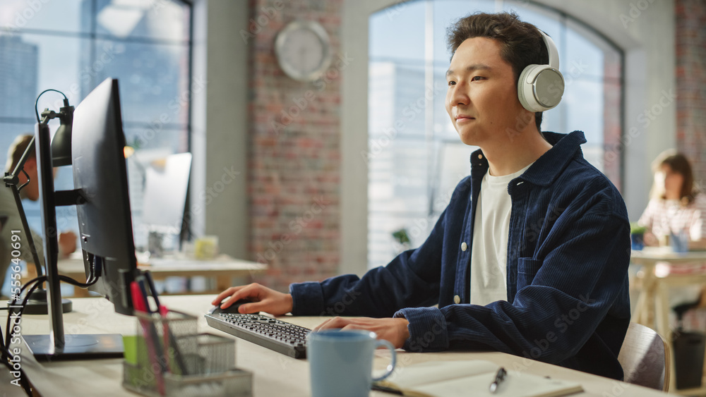Poster portrait of asian creative focused man smiles while using computer and headphones in bright modern o
