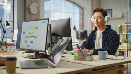 Portrait of an Asian Man Smiling While Using Computer and Headphones in Bright Modern Office. Communications Coordinator Planning a Project While Listening to Podcast.