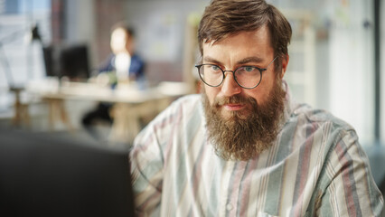 Fototapeta premium Portrait of Young Caucasian Bearded Man with Glasses Focused and Working on Computer in a Bright Spacious Office. Male Administrator Smiling after Successfully Finishing a Task.