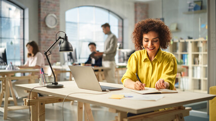 Portrait of Biracial Active Young Woman Working on Laptop in Busy Office. Biracial Female Manager smiling while Taking Notes one a Notebook From Her Laptop . People Using Computers in the Background.