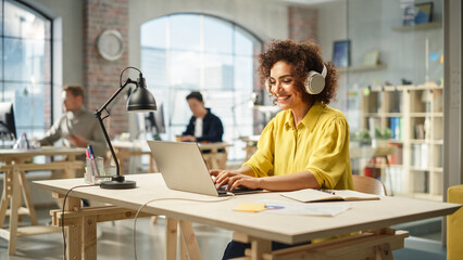 Portrait of Biracial Young Woman Smiling and Using Laptop in Bright Contemporary Office. Client Advisor Answering Customer Support Through Headphones and Typing. Team Working in the Background