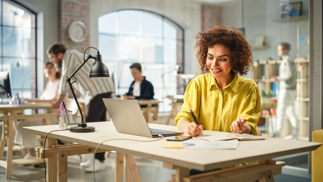 Portrait of Young Biracial Woman Taking Notes at Her Desk While Looking for References on the Laptop. Busy Employees Are Collaborating in the Background.