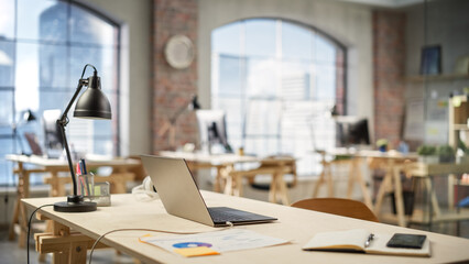 Wired Laptop on a Desk in a Bright Spacious Empty Workplace. Office Supplies Are on the Table. Big Windows with City Skyscrapers View in the Background.