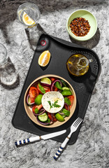 Greek salad in a restaurant serving on a black board with olive oil. The salad contains cucumbers, tomatoes, lime, cheese, onions, herbs, spices and lemon. Olive oil salad dressing.