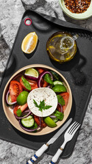 Greek salad in a restaurant serving on a black board with olive oil. The salad contains cucumbers, tomatoes, lime, cheese, onions, herbs, spices and lemon. Olive oil salad dressing.