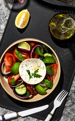 Greek salad in a restaurant serving on a black board with olive oil. The salad contains cucumbers, tomatoes, lime, cheese, onions, herbs, spices and lemon. Olive oil salad dressing.