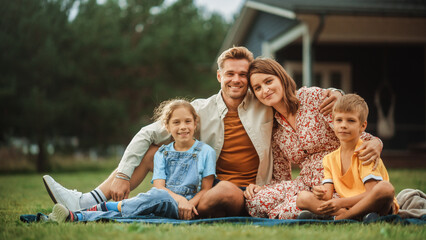 Portrait of a Happy Young Family with Kids Sitting on a Lawn in Their Front Yard on a Warm Summer Day at Home on a Picnic. Cheerful People Looking at Camera and Smile.