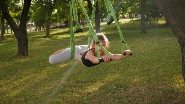 A little active teenage girl practices aerial yoga while lying in a green hammock hanging from a tree in the park. A child does yoga outdoors.