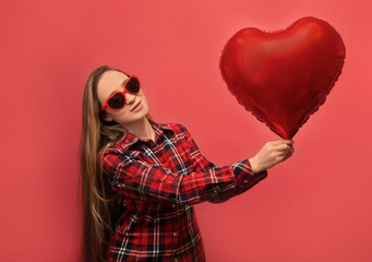 Saint Valentines Day and love concept: young woman with long hair holding heart shaped air balloon, wearing heart party glasses and casual checkered plaid shirt isolated on colored pink background.