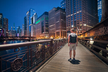 Rear view of man with backpack while walking on bridge and looking around. Illuminated city with skyscrapers at twilight. Chicago, United States..