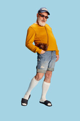 Portrait of stylish, senior, bearded man in casual clothes with cap, sunglasses and vintage camera posing over blue background