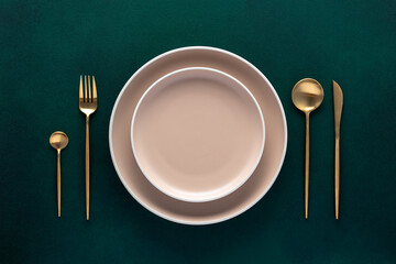 Festive place setting with beige dishes. Empty plate and gold cutlery on dark green background....