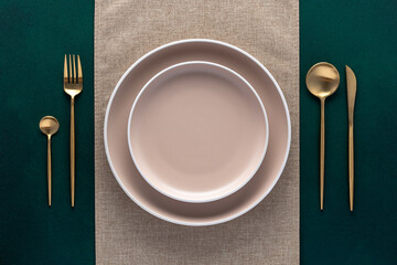 Festive place setting with beige napkin. Empty plates and gold cutlery on dark green background. Top view. Dining table in luxury restaurant. Card or menu template, flat design. Tableware, crockery. - obrazy, fototapety, plakaty