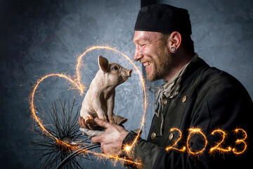 Schornsteinfeger in schwarzer Arbeitskleidung hält Glücksschwein in der Hand, Herz und Jahreszahl 2023 mit strahlenden Wunderkerzen geschrieben.