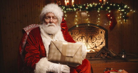 Authentic santa claus sitting in his rocker near fireplace, looking at camera and offering a christmas gift - christmas spirit, holidays and celebrations concept 