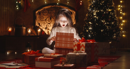 Obraz na płótnie Canvas Little caucasian girl sitting near christmas tree in decorated room and opening her gift with something special - holidays and celebrations, christmas spirit concept close up 