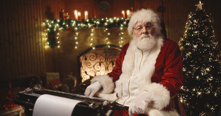 Joyful santa claus sitting at his typewriter, looking at camera and playfully winking in specially decorated room - christmas spirit, holidays and celebrations concept 