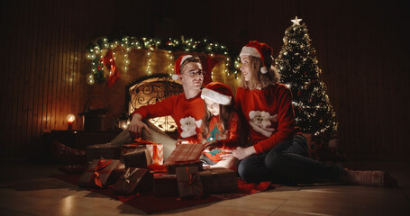 Caucasian family of three sitting in decorated room near christmas tree, little girl opening her christmas gift with something special - christmas spirit, togetherness concept 