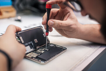 Workplace top view, close-up. Repairman disassembles smartphone with screwdriver in an electronics...