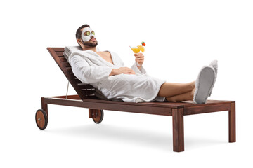 Man in a bathrobe with a face mask and cucumber on eyes resting on a deck lounge chair with a cocktail