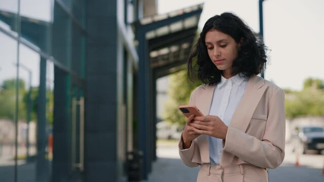 Beautiful curly smiling young businesswoman in formal outfit standing near the business centre using smartphone. People and technology business concept