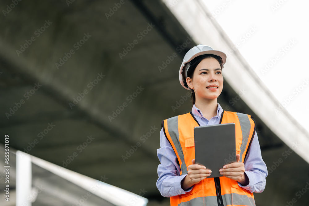 Wall mural Asian engineer worker woman or architect looking construction with tablet wear white safety helmet in construction site. Standing at highway concrete road site. Progress planning of highway bridge.