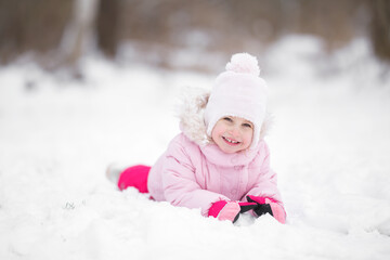 Fototapeta na wymiar Smiling beautiful little girl lying down on white snow at city park. Cute 3 years old toddler enjoying cold winter day. Front view. Closeup. Posing and looking at camera.