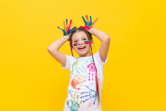 A dabbling girl stained in paints shows horns on a yellow isolated background. The child is playing with colorful paints. Courses for the development of children's creativity. Happy childhood games.
