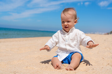 Little happy barefoot boy child sits on the beach and plays with sand on a sunny day.