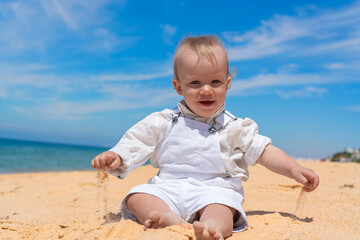 Little happy barefoot boy child sits on the beach and plays with sand on a sunny day.
