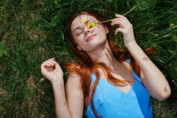 Woman smile with teeth lying on the green grass in a blue dress in the spring sunshine with yellow flowers, happiness, red long hair