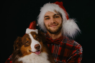 Dog and human with Santa Claus hats on their heads. Studio shot. Concept of pet celebrating Merry Christmas with male owner. Young Caucasian man with beard hugs Australian Shepherd.