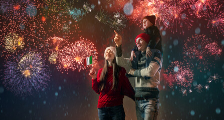 Mom and dad with their son on their shoulders in warm clothes and in a Santa hat. The family celebrates the new year looking at the fireworks outside
