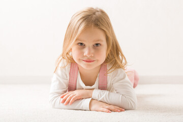 Smiling beautiful little girl lying down on belly on light beige home carpet. Cute 3 years old...