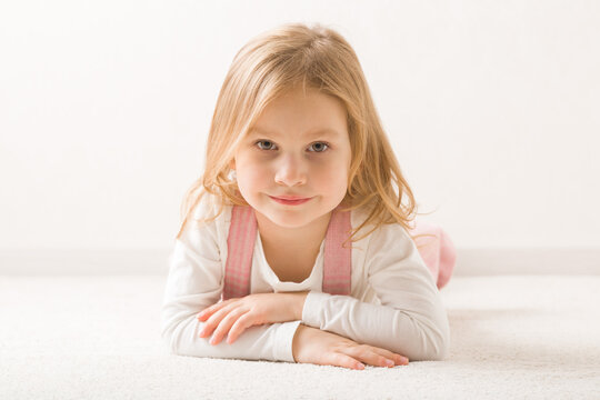 Smiling beautiful little girl lying down on belly on light beige home carpet. Cute 3 years old toddler. Front view. Closeup.