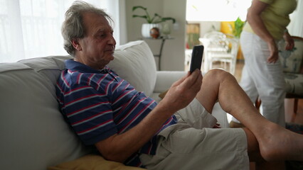 Obraz premium Senior man holding phone speaks in video with smartphone. Casual authentic older mature person using modern technology device in living room couch