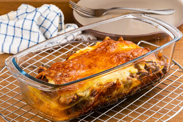 High angle view of freshly baked homemade lasagna in transparent tray on cooling rack with brown ceramic dish and metal fork on wooden table.
