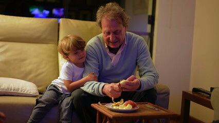 Grandson and grandfather eating apple together bonding at living room sofa at night. Grandparent family generational interaction