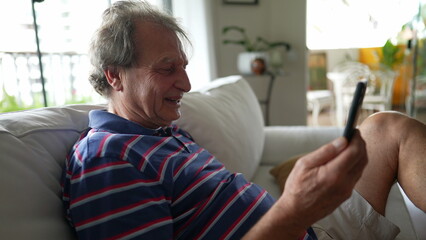 One happy senior man laughing and smiling while speaking with family member on cellphone device seated on sofa couch at living room home