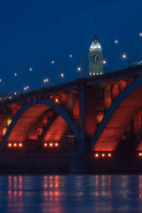 Obraz premium Evening cityscape with red illuminated of Communal Bridge and Clock tower in Krasnoyarsk, Russia. View of the Yenisey River at dusk. Blurred reflection of lighting lights in the rippled water surface